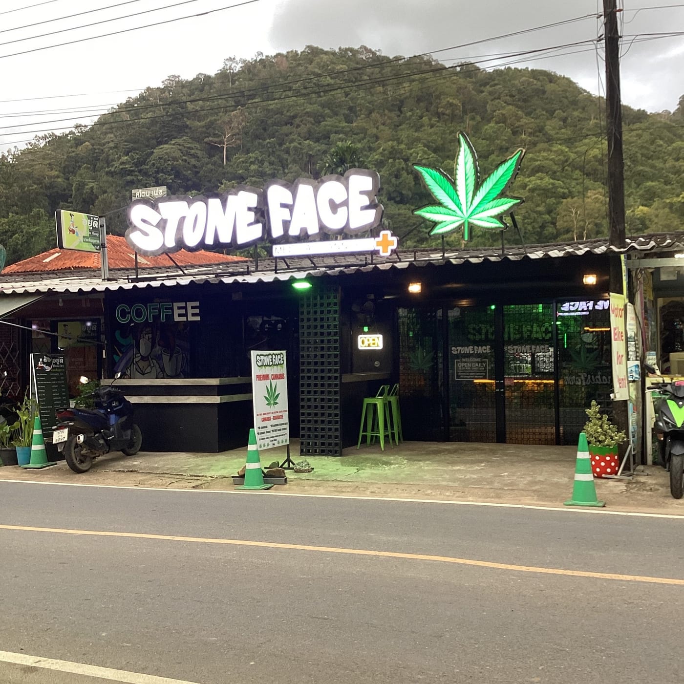 Stone Face from the road in daylight — the lit sign and green neon leaf above an open-fronted shop, COFFEE painted on the left wall, forested hills behind